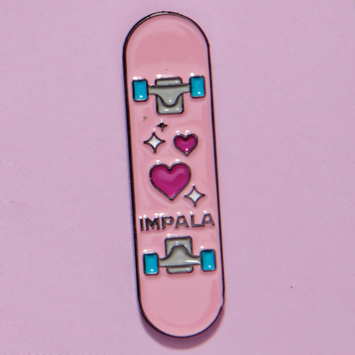 Pink skateboard-deck enamel pin with blue details, hearts, and small sparkles
